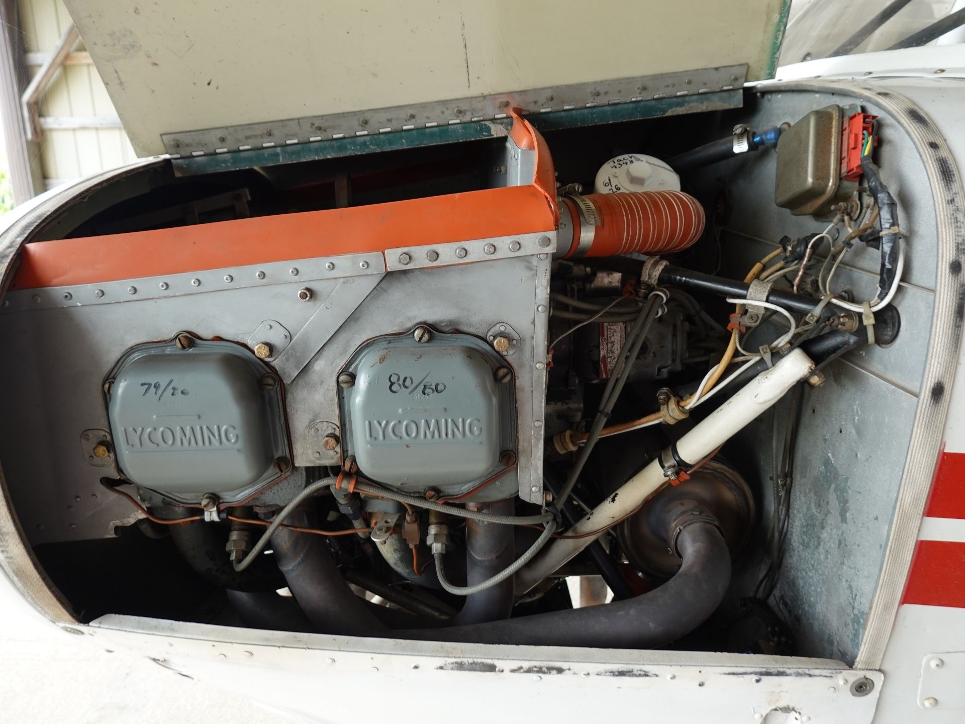 Open engine cowling of a small aircraft showing Lycoming engine cylinders and wiring, with orange ducting and various hoses and components visible inside the nacelle.