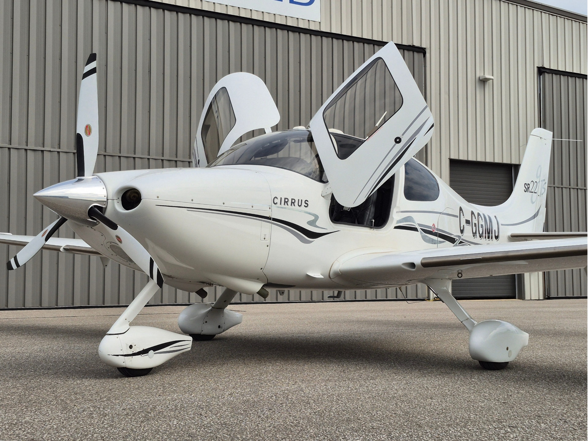 White Cirrus SR22 airplane with gull wing doors open parked on tarmac in front of a corrugated metal hangar