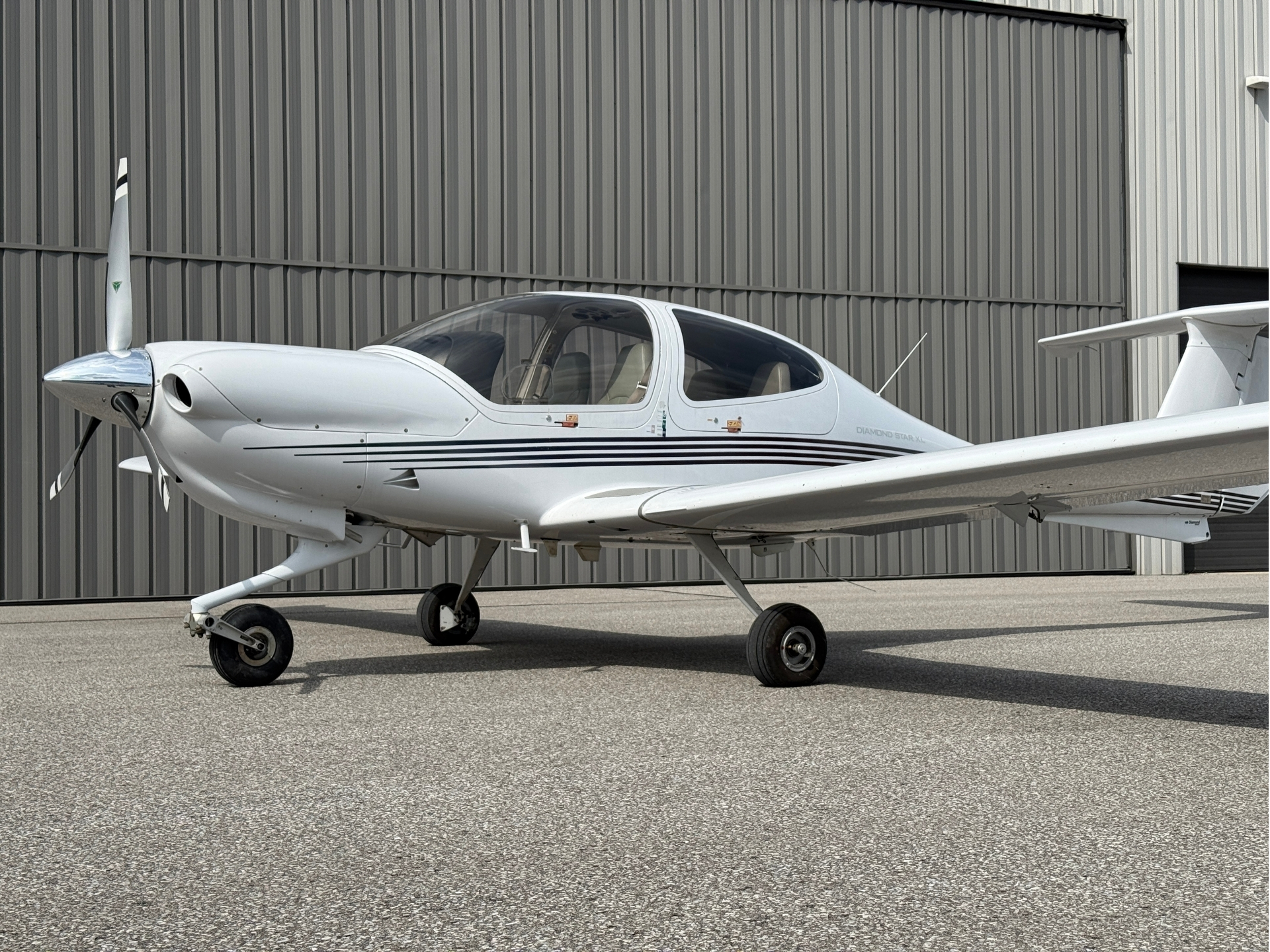 White single-engine propeller airplane parked on tarmac in front of a corrugated metal hangar.