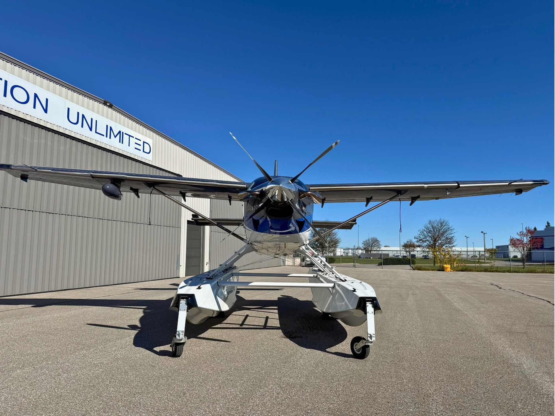 Front view of a silver blue floatplane on its landing gear parked on a tarmac beside a hangar with UNLIMITED signage