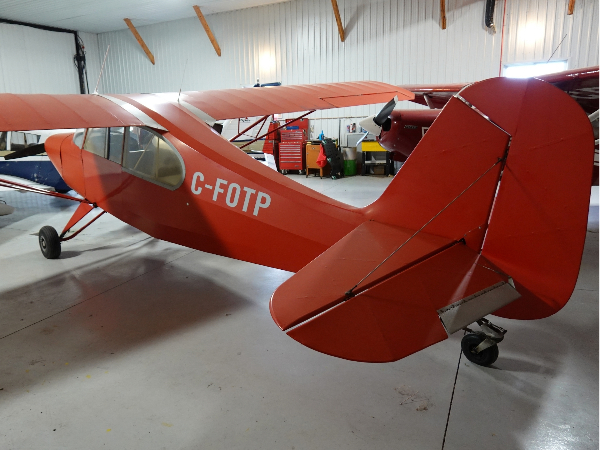 Red vintage single engine airplane in a hangar registration C FOTP with a visible cockpit canopy and prop at the nose workshop tools in the background