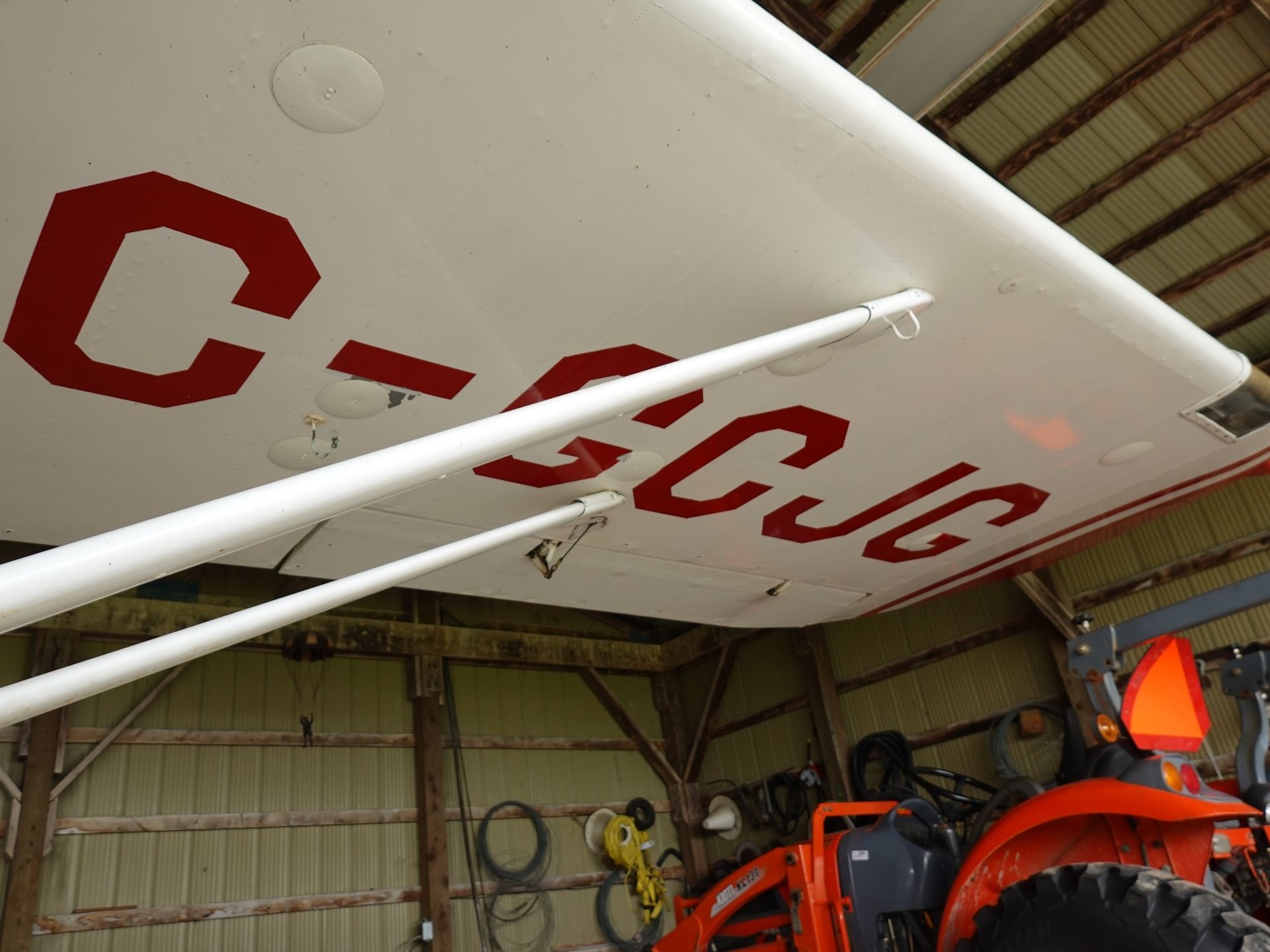 Close-up of a light-gray airplane wing with large red registration letters, inside a hangar with orange maintenance equipment nearby.