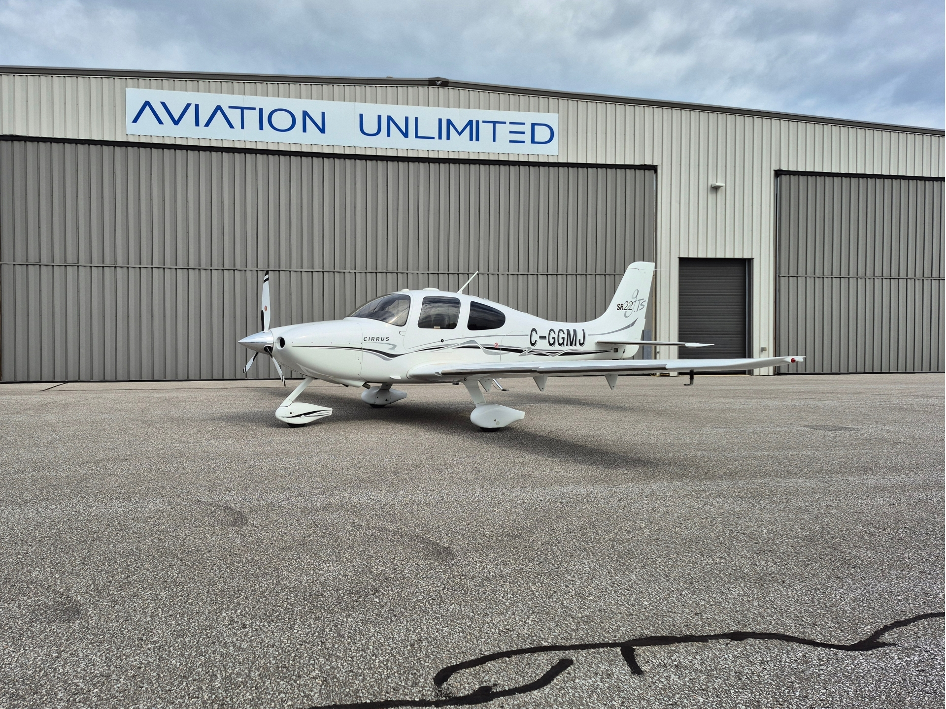 White Cirrus small airplane parked on the tarmac in front of a grey hangar with a blue AVIATION UNLIMITED sign above it