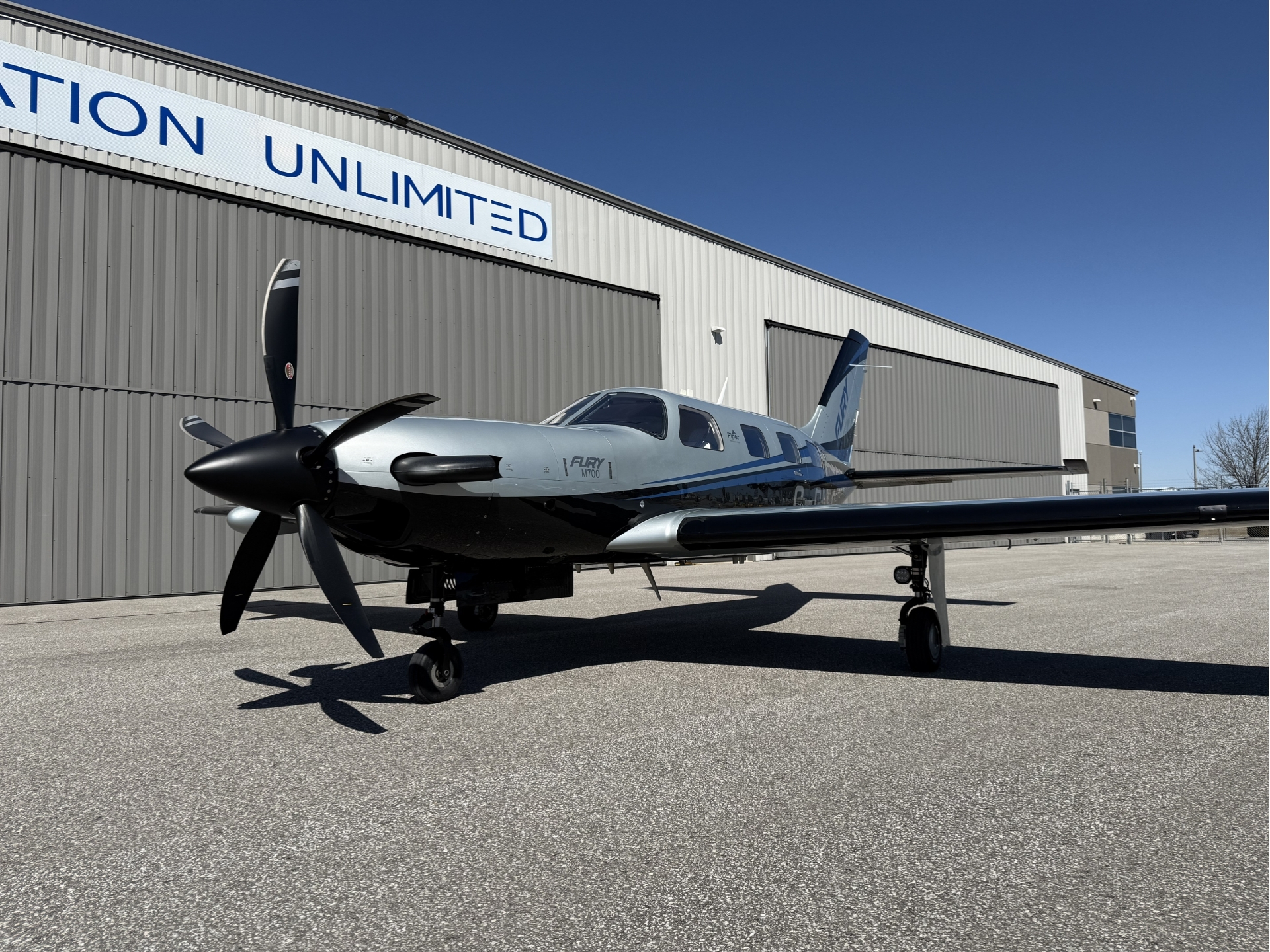Silver twin engine propeller airplane parked on the tarmac beside a large gray hangar with a blue sky overhead fuselage bears the name Fury M700