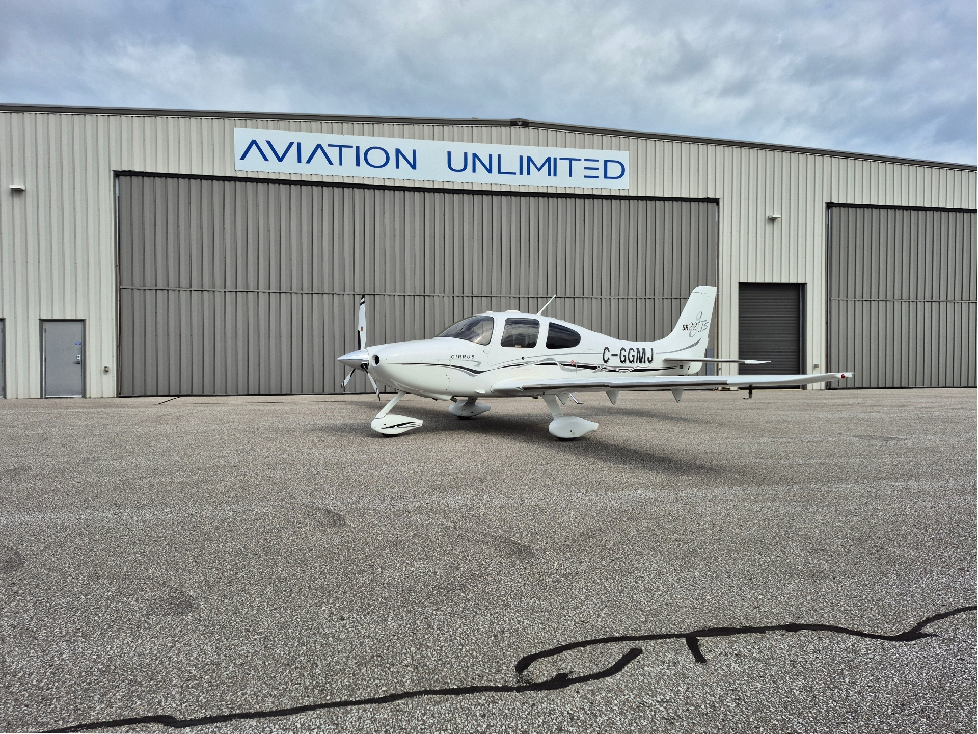 White Cirrus SR22 airplane parked on a tarmac in front of a gray hangar with a blue AVIATION UNLIMITED sign above the doors