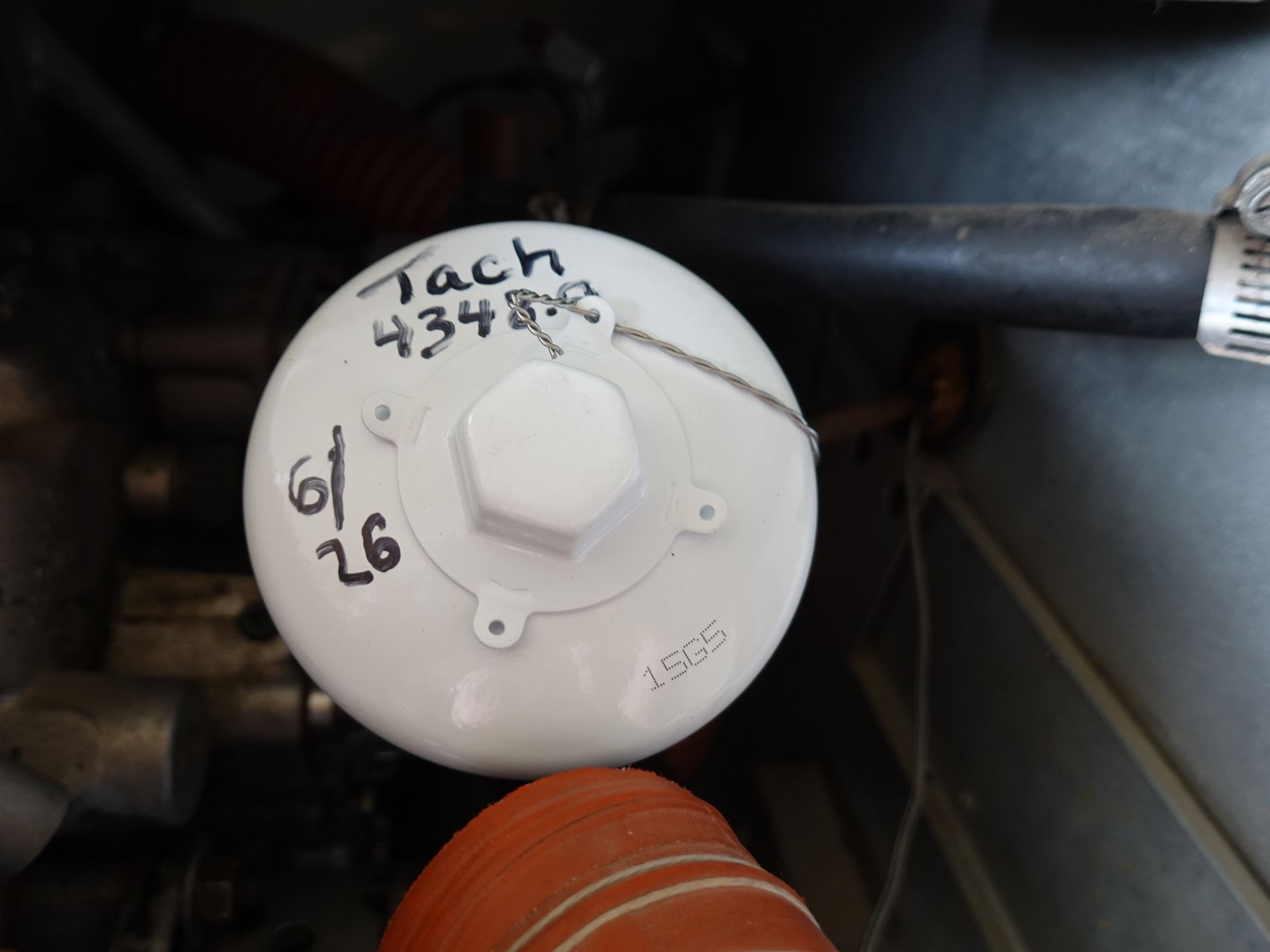 Close-up of a white circular engine reservoir cap with handwritten numbers and a braided wire on top, in a dim engine bay with an orange hose nearby.