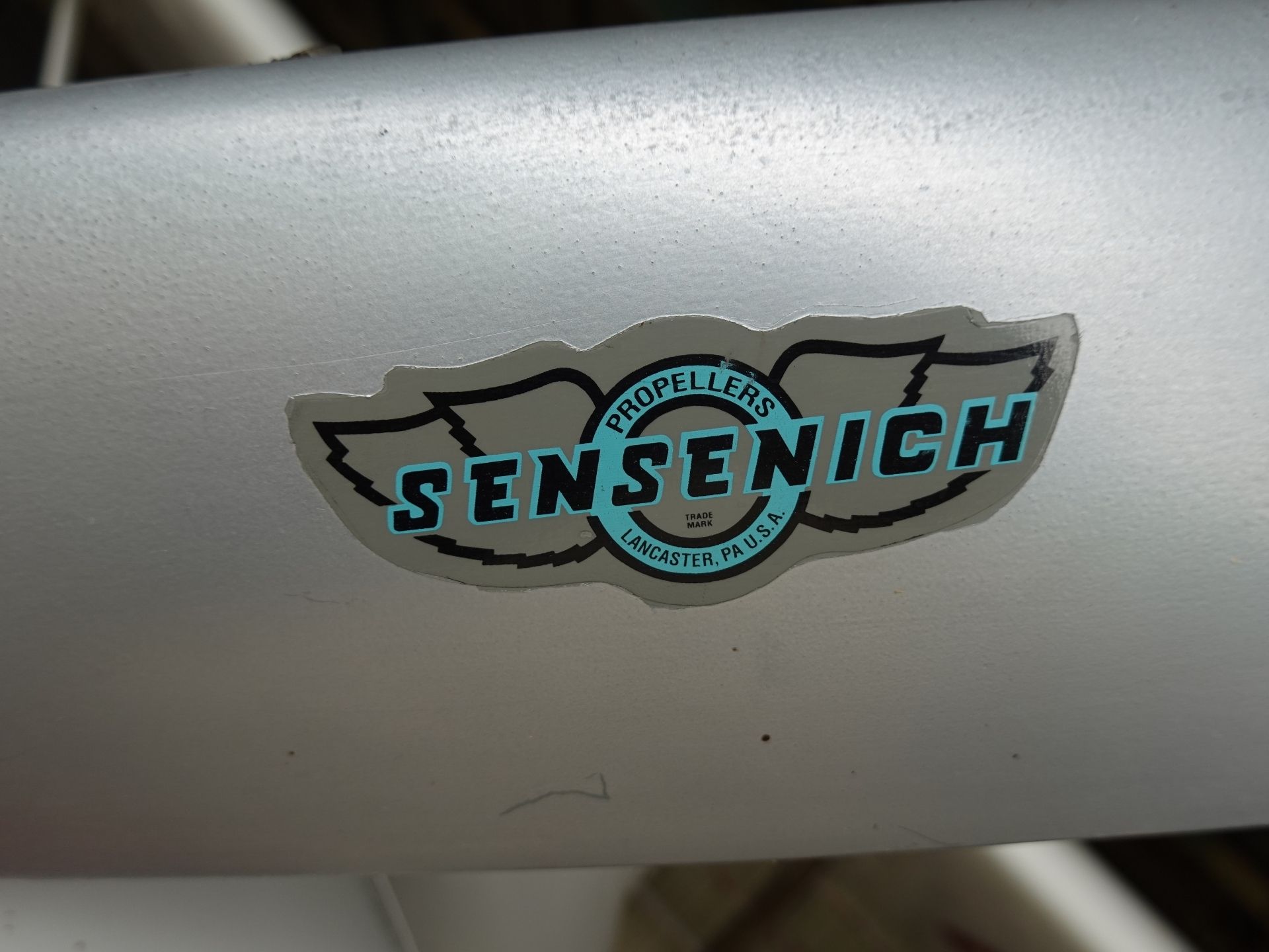 Sticker on a gray metal surface reading 'Sensenich Propellers' with a winged logo and 'Lancaster, PA USA' text. Alternative: close-up of a Sensenich Propellers decal on a light metal surface.