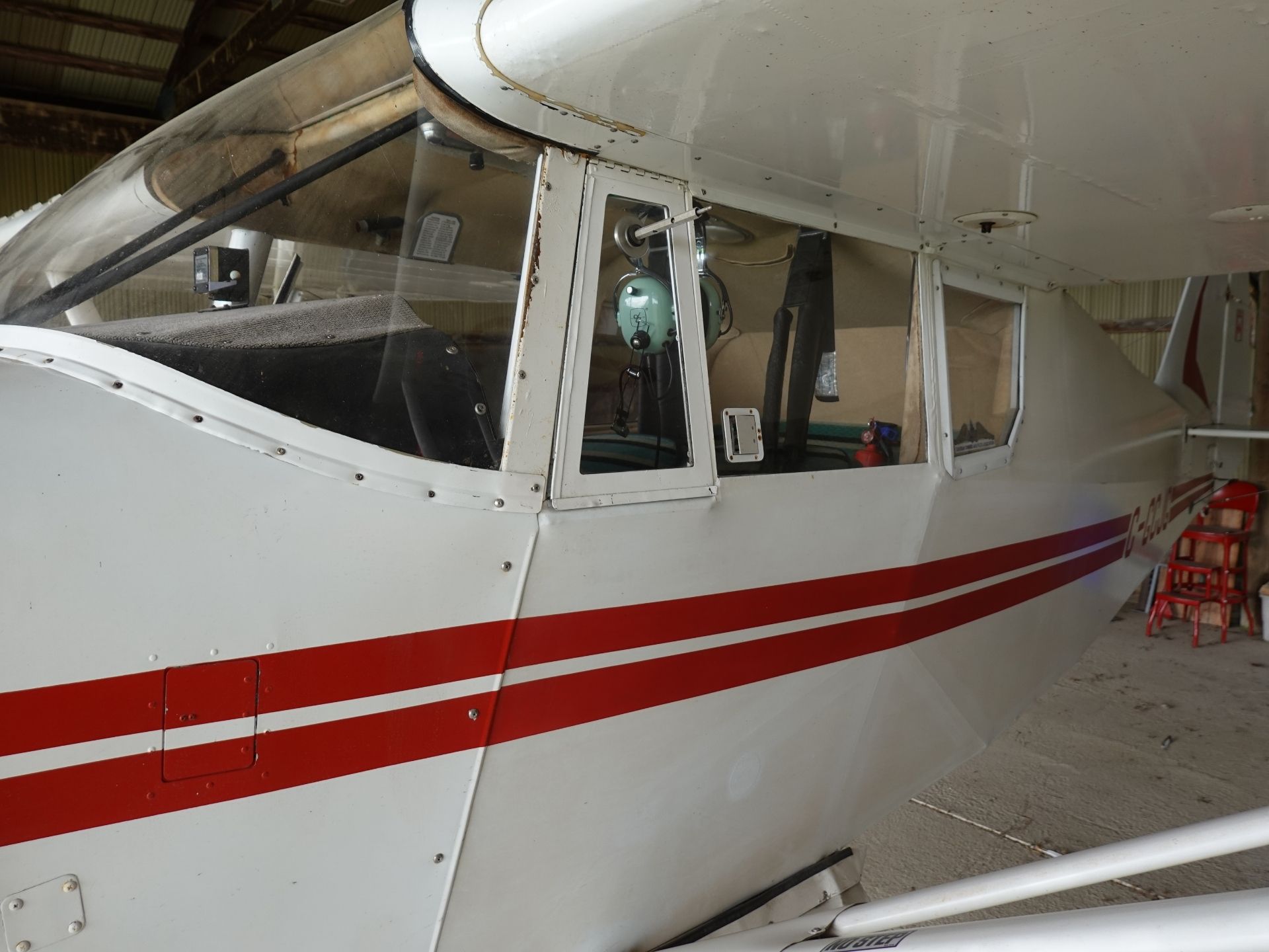 Small white airplane in a hangar with a red stripe along the fuselage, cockpit windows visible and a green headset hanging inside the cockpit.