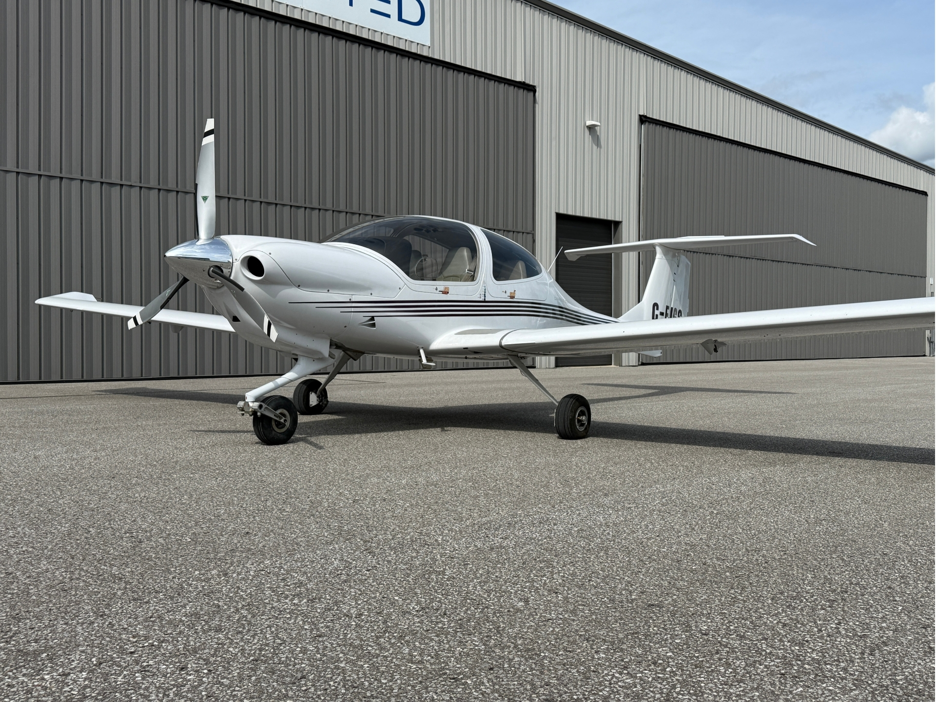 White single-engine airplane parked on a tarmac in front of a gray metal hangar, with propeller at the nose and clear cockpit canopy.