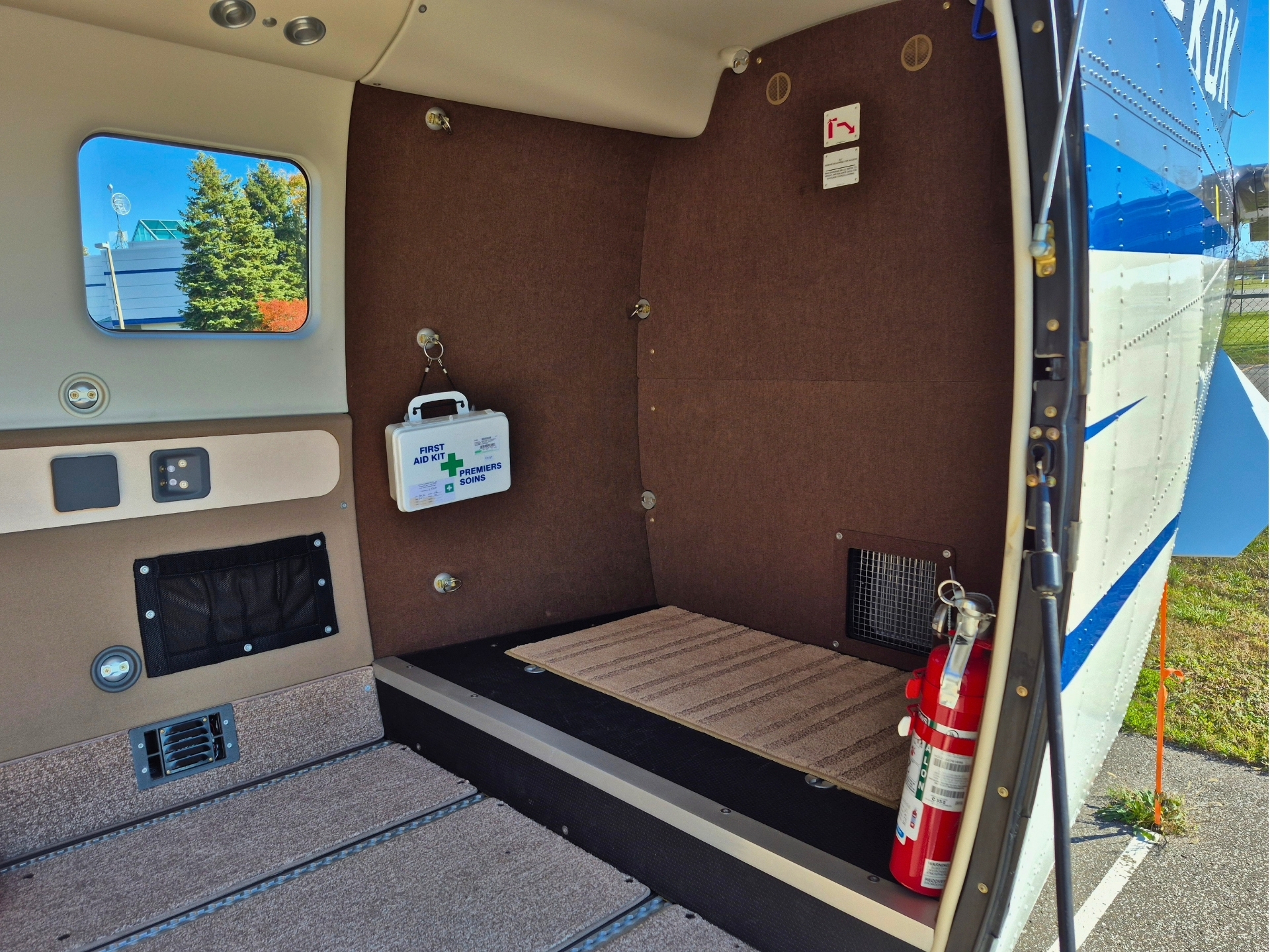 Interior of a small airplanes cargo area with brown fabric walls a hanging first aid kit and a red fire extinguisher near the open doorway window shows blue sky and trees outside