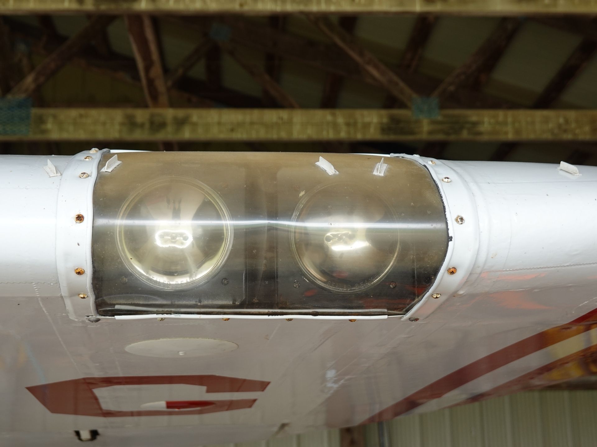 Underbelly of an aircraft with two round windows in a glass panel, riveted aluminum skin, inside a hangar with wooden beams above.