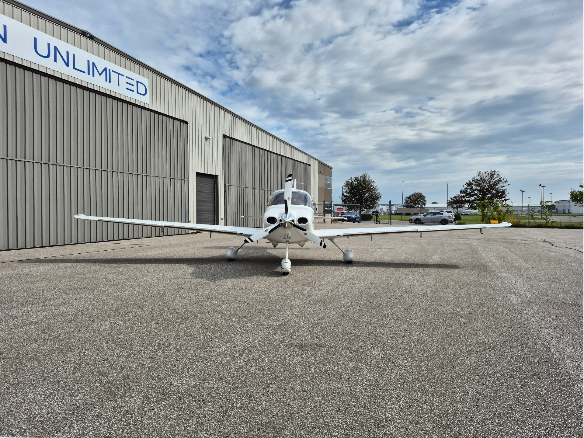 Small propeller plane parked on a tarmac in front of a large hangar with UNLIMITED signage under a cloudy blue sky