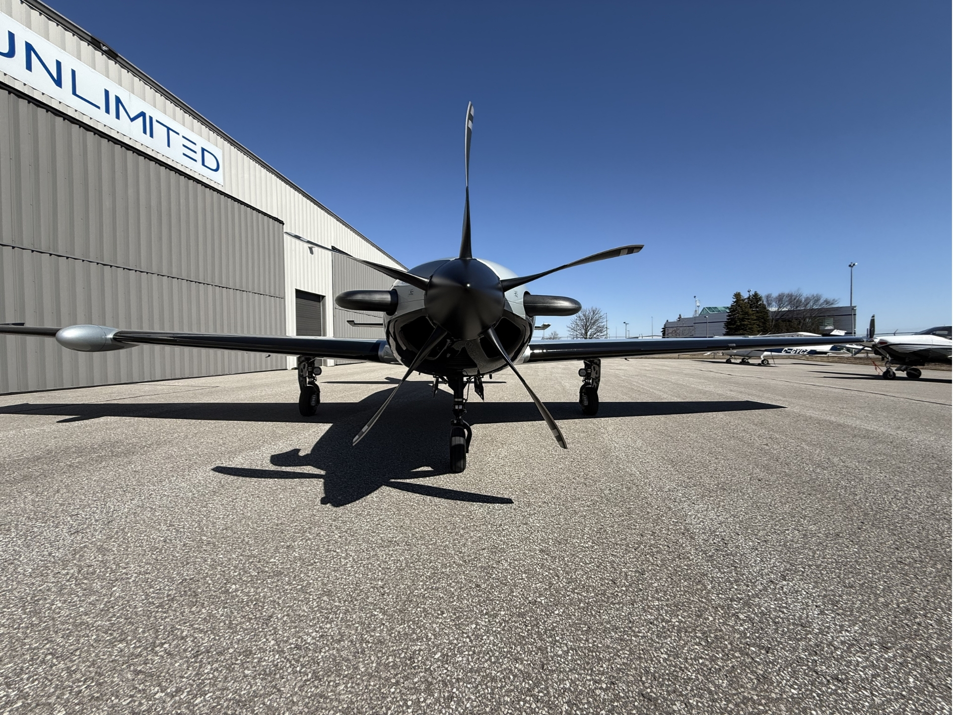Front view of a small propeller airplane parked on a tarmac in front of a hangar with a clear blue sky above