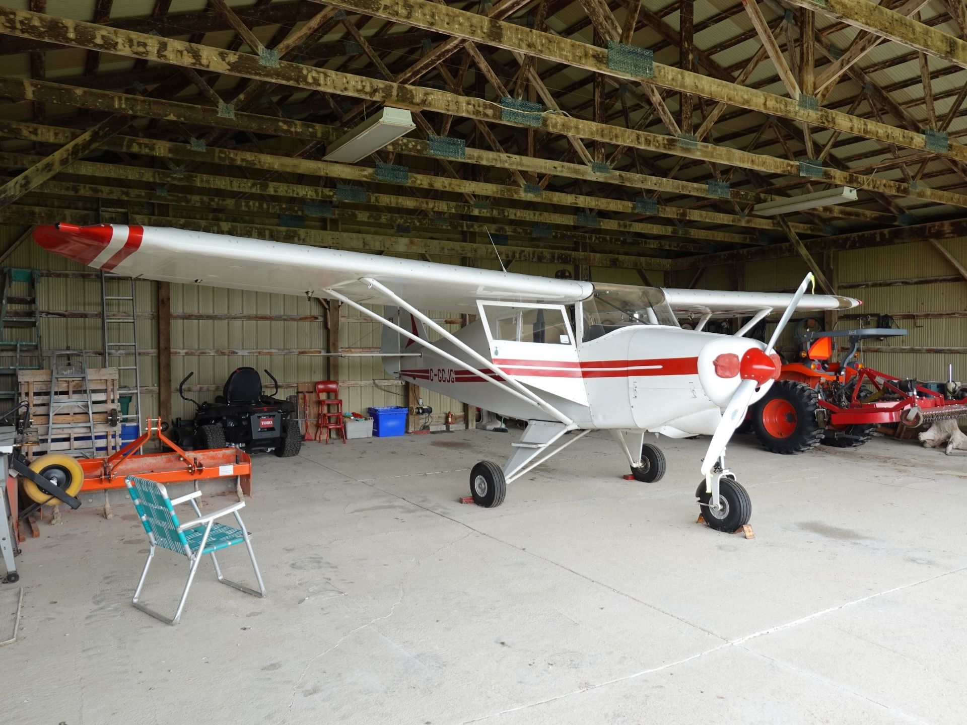 White single-engine airplane with red accents parked inside a wooden-beamed hangar, surrounded by tools, ladders, and equipment.