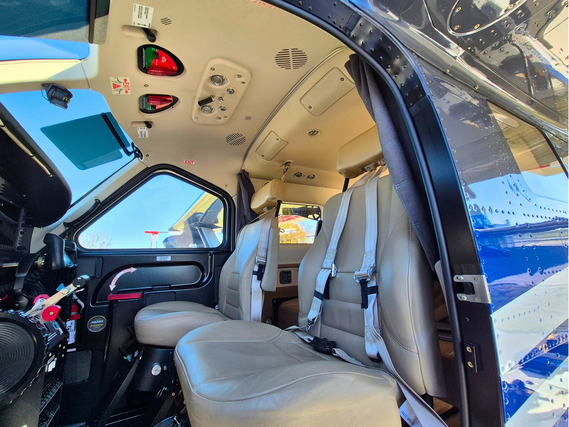 Interior of a helicopter cockpit with beige seats and flight controls door open to blue exterior