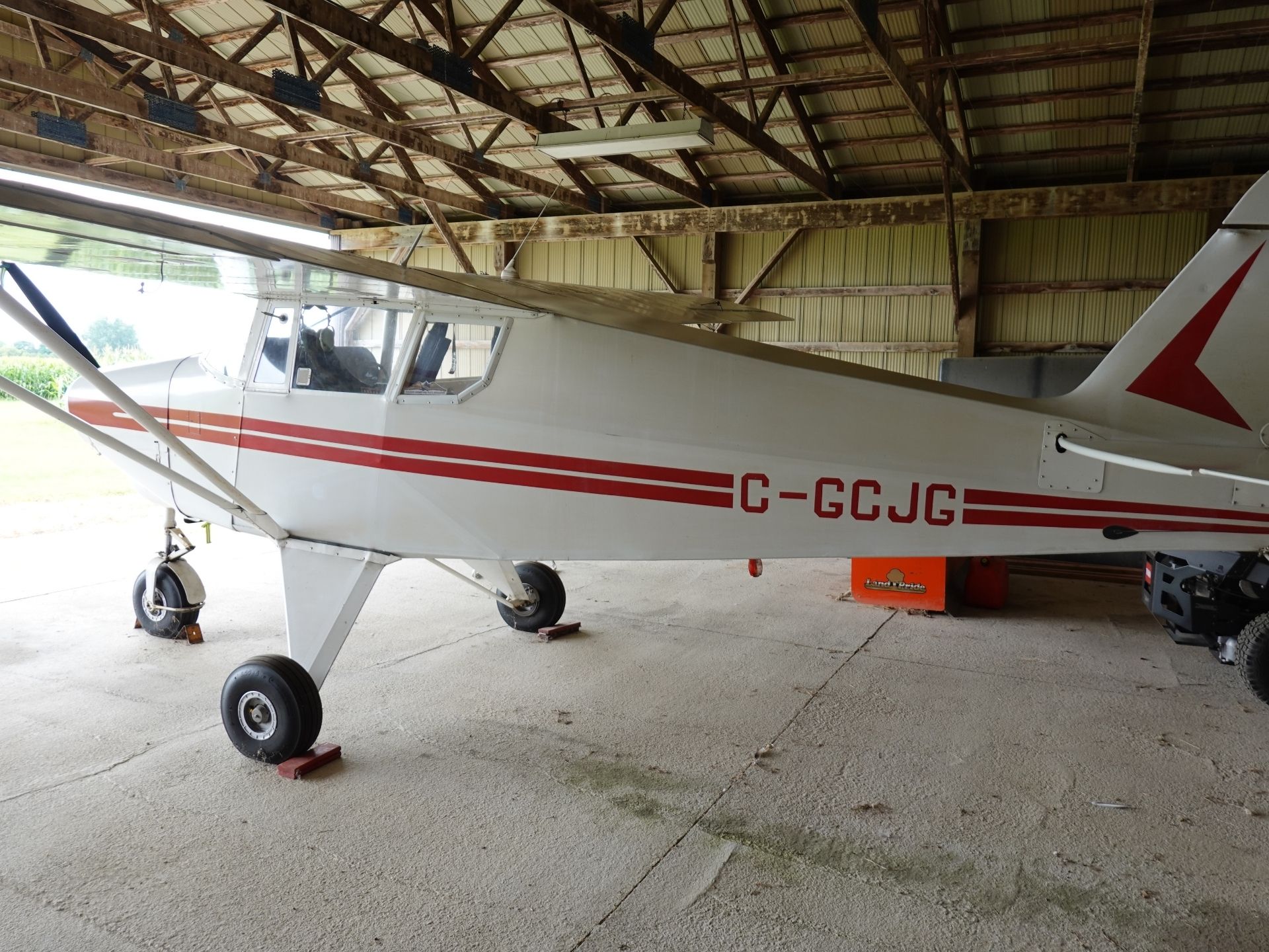 Small white single-engine airplane with red stripes in a hangar; nose, cockpit, and landing gear visible. (Registration C-GCJG)