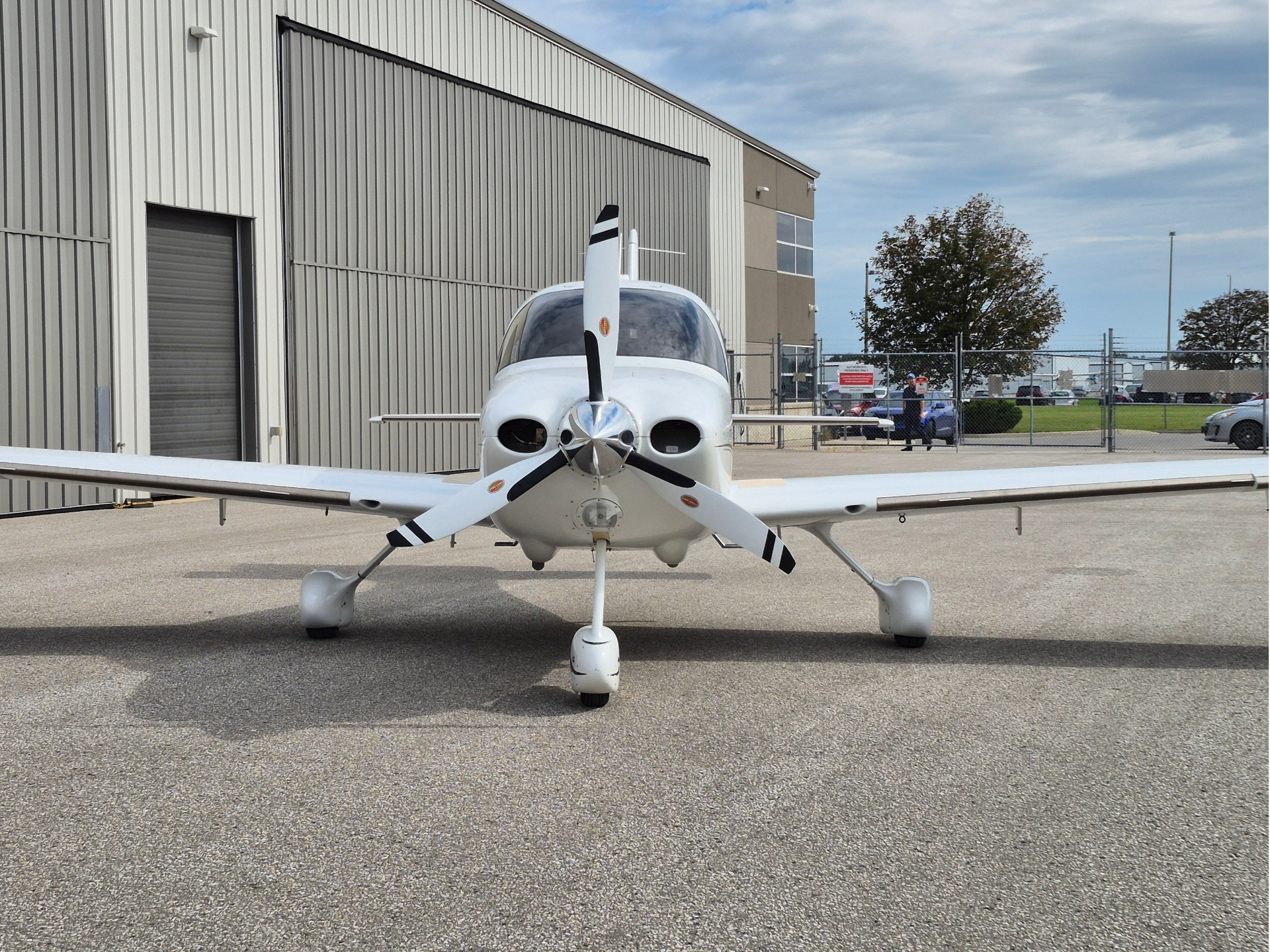 Front view of a white single engine propeller plane parked on a tarmac with a hangar in the background on a clear day