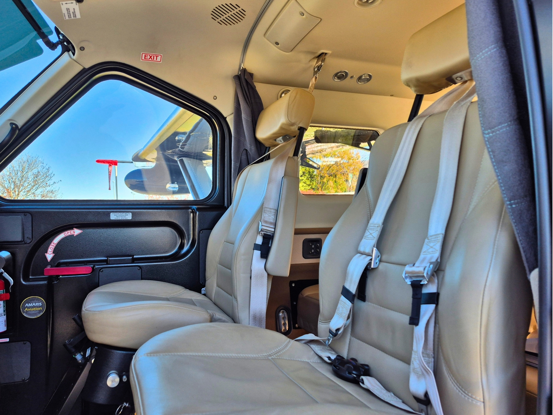 Inside a small aircraft cockpit beige seats with buckled seatbelts and a window view of blue sky outside