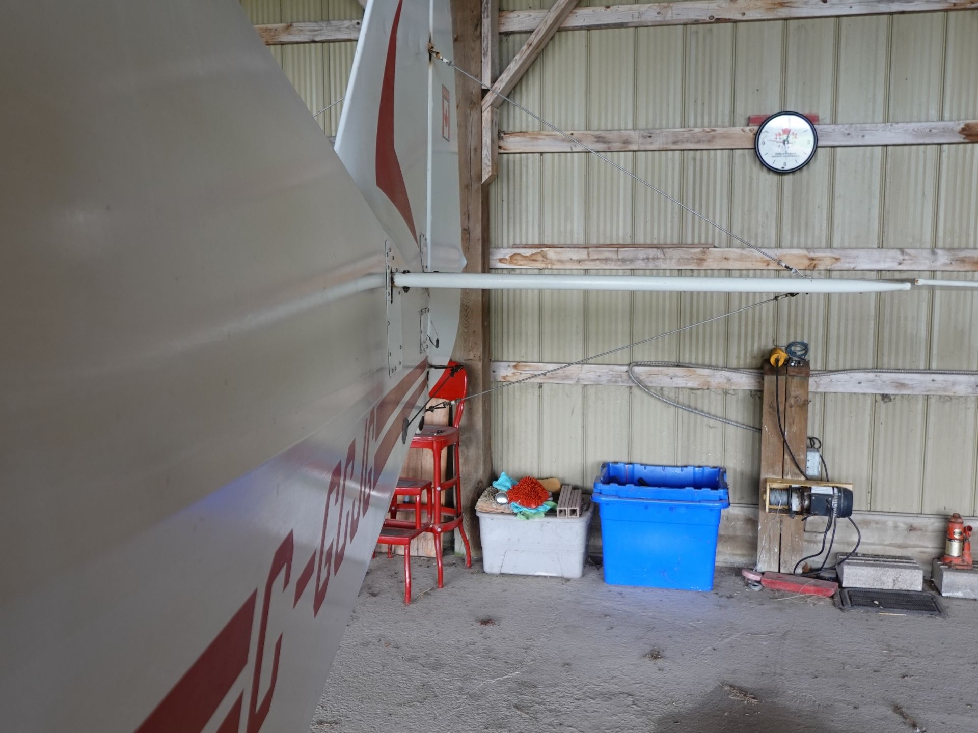 Side of a aircraft fuselage inside a hangar, with red stools, blue bin, and tools against a wooden beam wall. Clock on the corrugated metal wall.