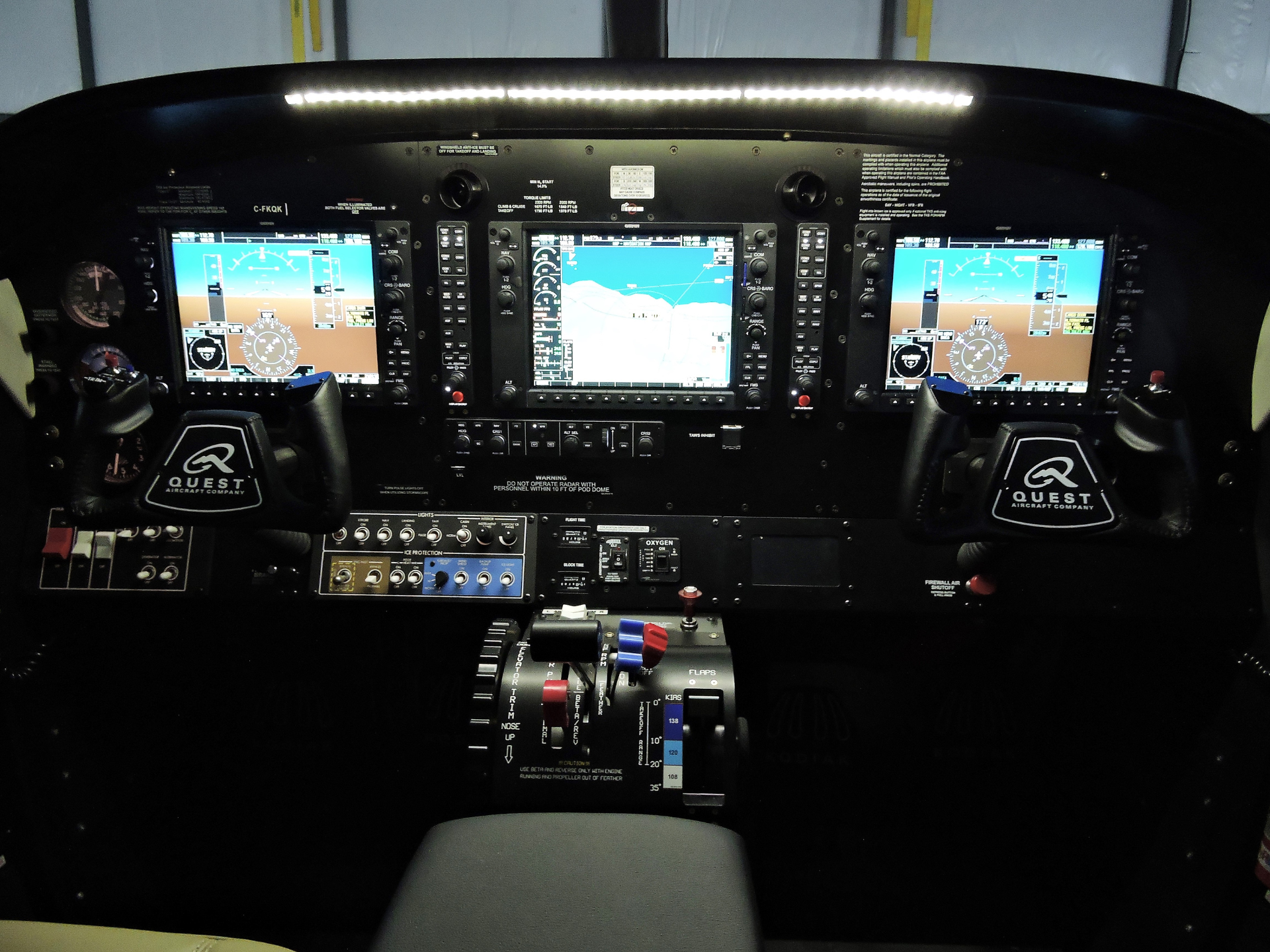 Cockpit of a small aircraft with three large avionics displays center console and throttle levers Quest Aircraft branding visible on yokes