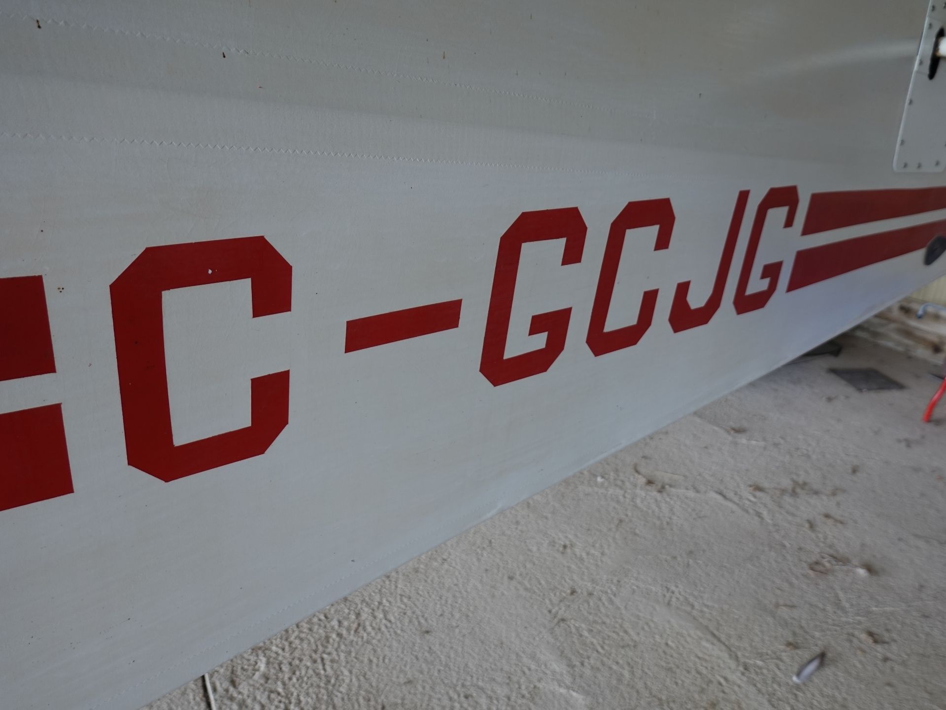 Side view of a light aircraft fuselage with large red block letters along the white body, photographed at ground level.