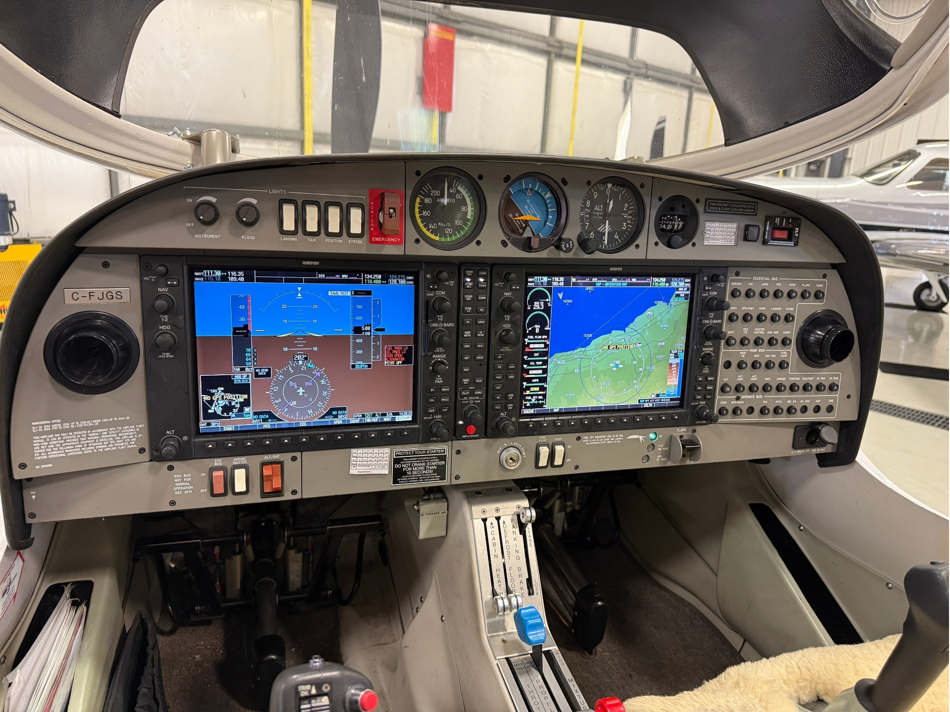 Close-up of a small aircraft cockpit showing Garmin flight displays, engine gauges, and control panels in a hangar setting.