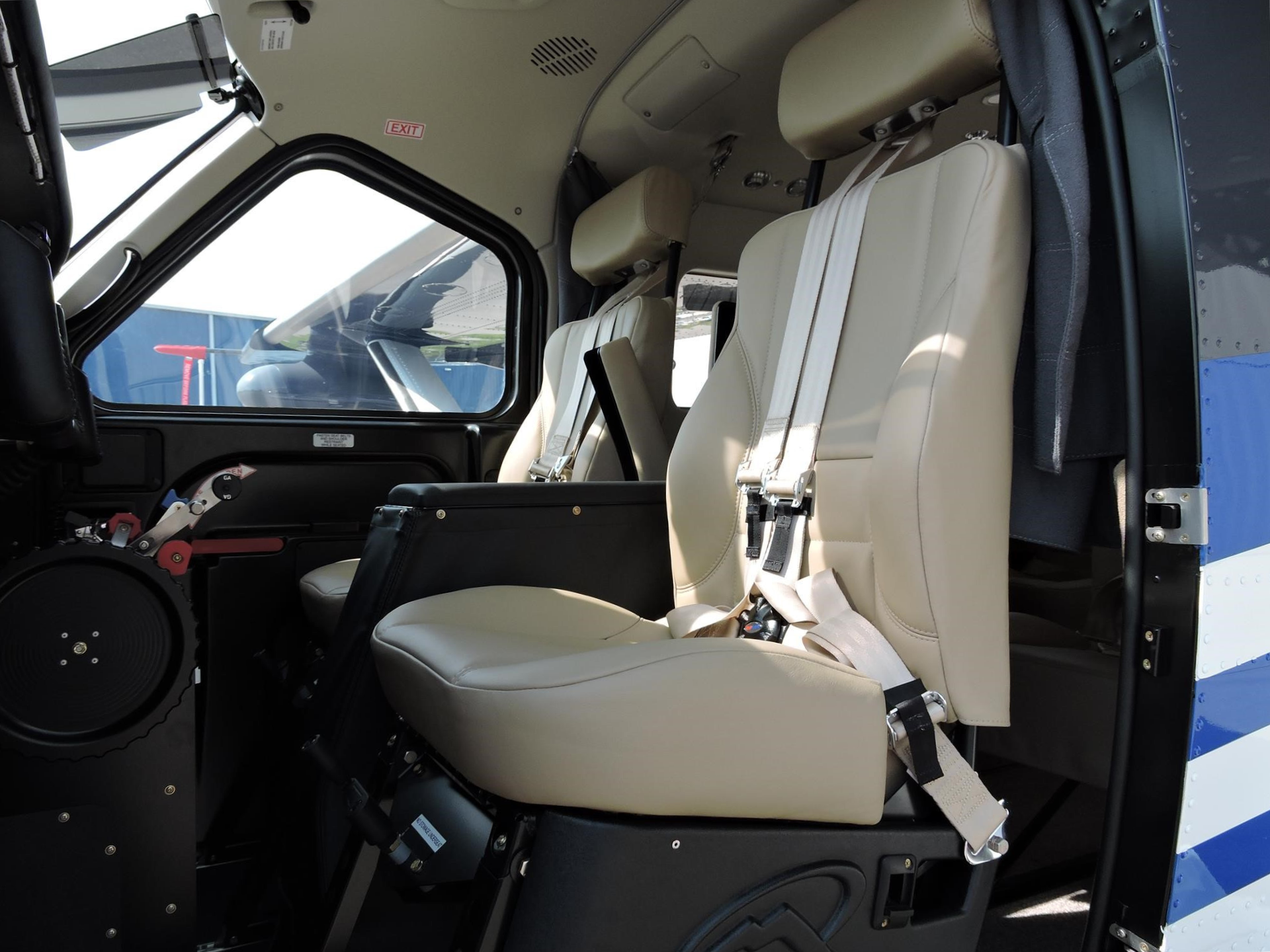 Interior of a small helicopter cabin with beige leather seats and shoulder harnesses exposed cockpit controls nearby