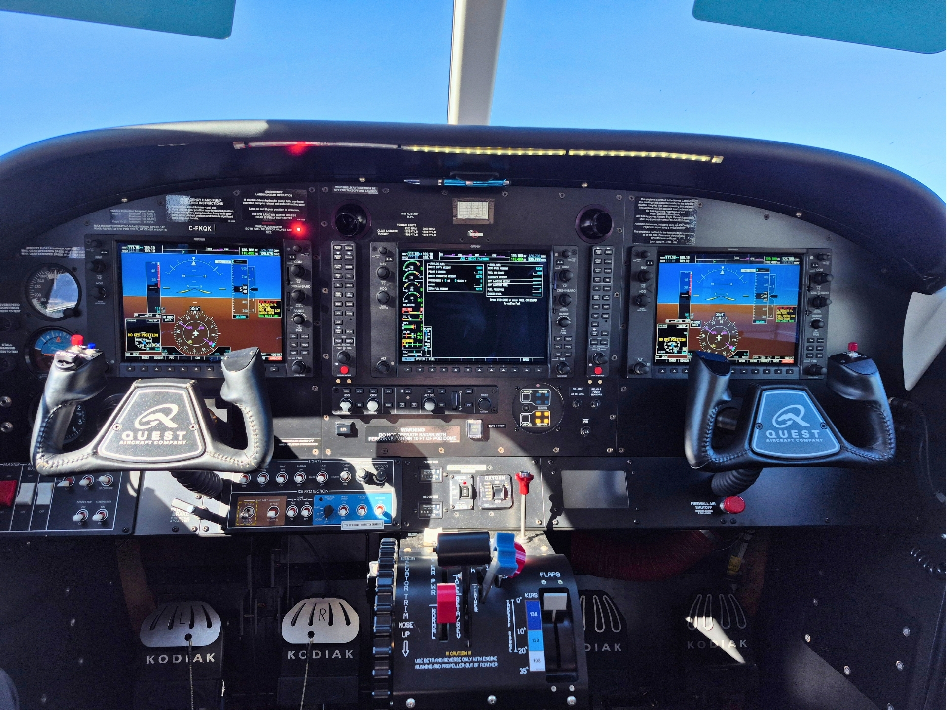 Front cockpit view of a glass cockpit with three large displays and central flight controls labeled Kodiak showing flight data and navigation
