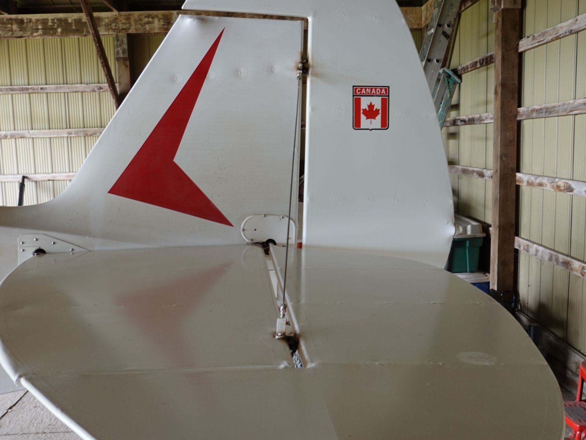 Rear fuselage and tail of a small white airplane in a storage hangar, featuring a red chevron and a Canada badge.